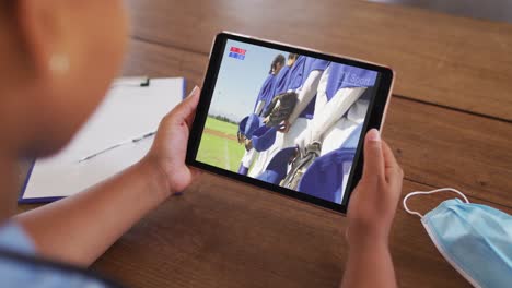 compuesto de una mujer sentada en una mesa con una máscara facial, viendo un juego de béisbol en la tableta