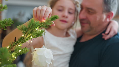 blurred view of caring dad carrying daughter in arms while she hangs beautiful white ornament on decorated christmas tree inside cozy festive home capturing emotional joyful family holiday moment