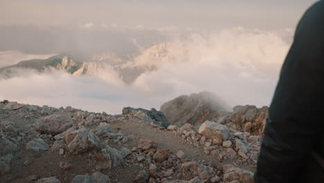 Young-man-with-a-backpack,-wearing-a-jacket,-head-covered-wiht-a-hood,-standing-on-a-peak-of-a-mountain-in-early-morning-hours,-watching-into-the-valley-where-view-is-partially-covered-with-clouds