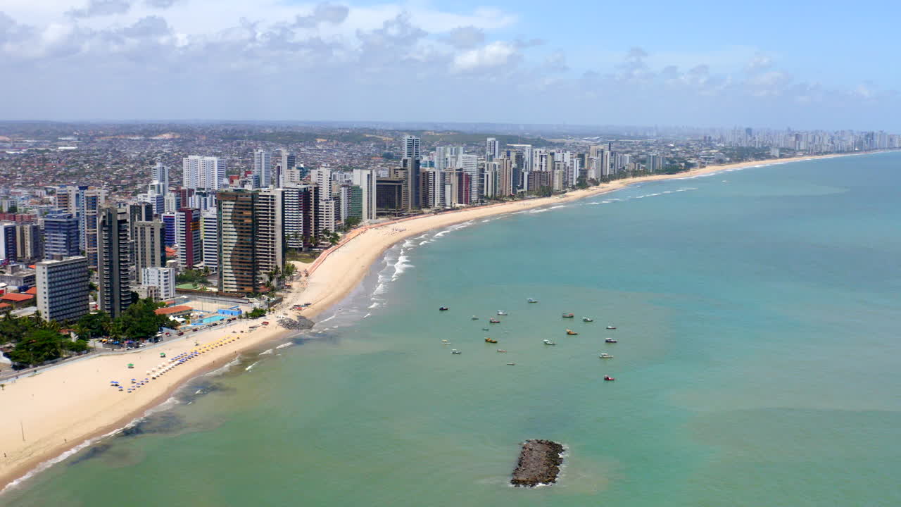 Drone footage of the vibrant coastline of Jaboatão dos Guararapes in Pernambuco Brazil showing green ocean waters sandy shores blue skies urban buildings summer holiday scene with boat and umbrella.