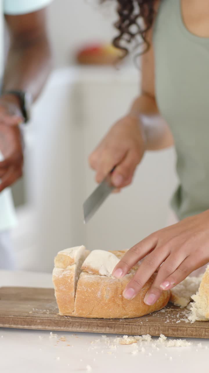 Vertical video: Woman steadying loaf and slicing with serrated knife in home cooking, man reaching