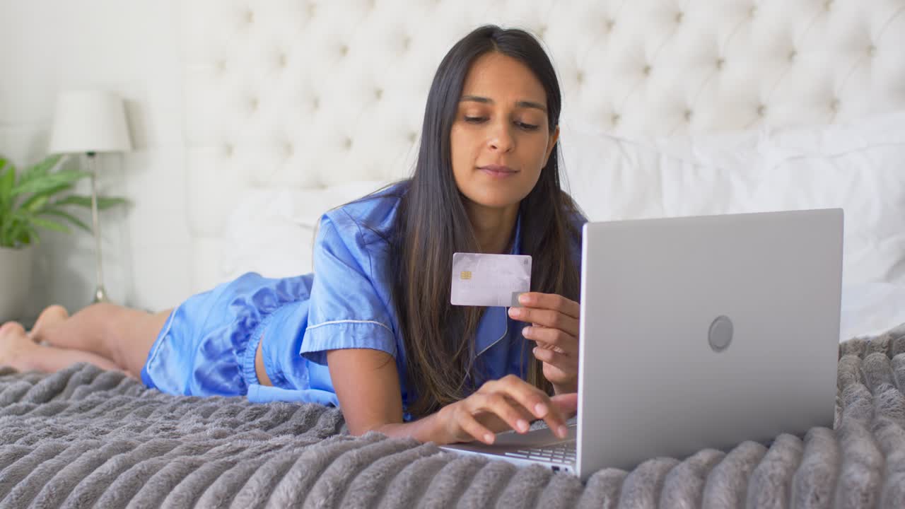 Mulher indiana deitada de bruços na cama, segurando um cartão de pagamento e digitando em um laptop prateado para pagar.