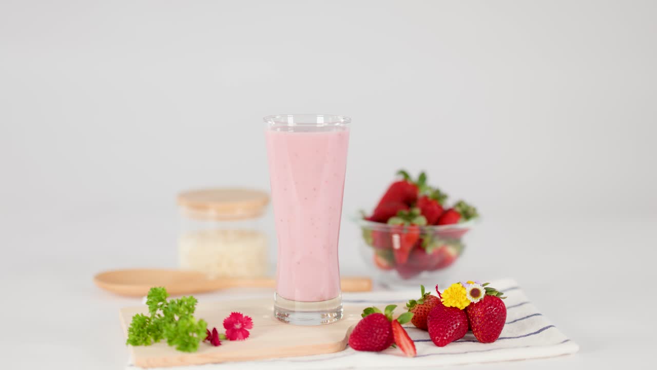 Una mano posiziona una fetta di fragola su un frappè rosa in uno studio luminoso, circondato da frutta fresca e fiori