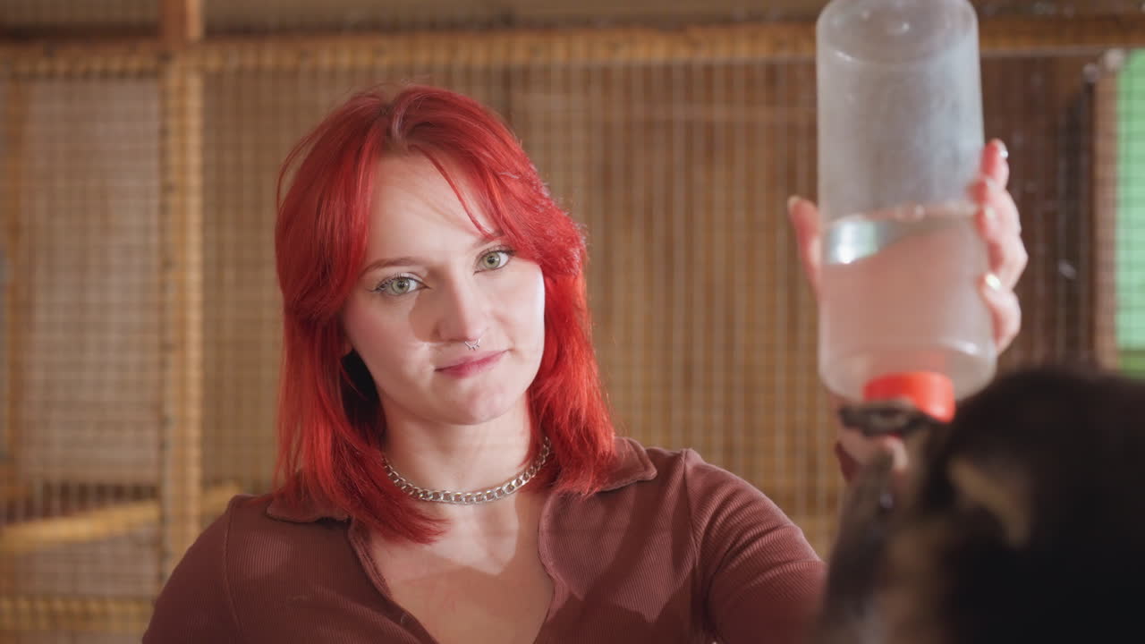 redhaired woman offering bottle to pet in shelter kennel gentle expression and steady gaze as volunteer feeds small animal, warm indoor light, visible necklace, patient interaction and bond building