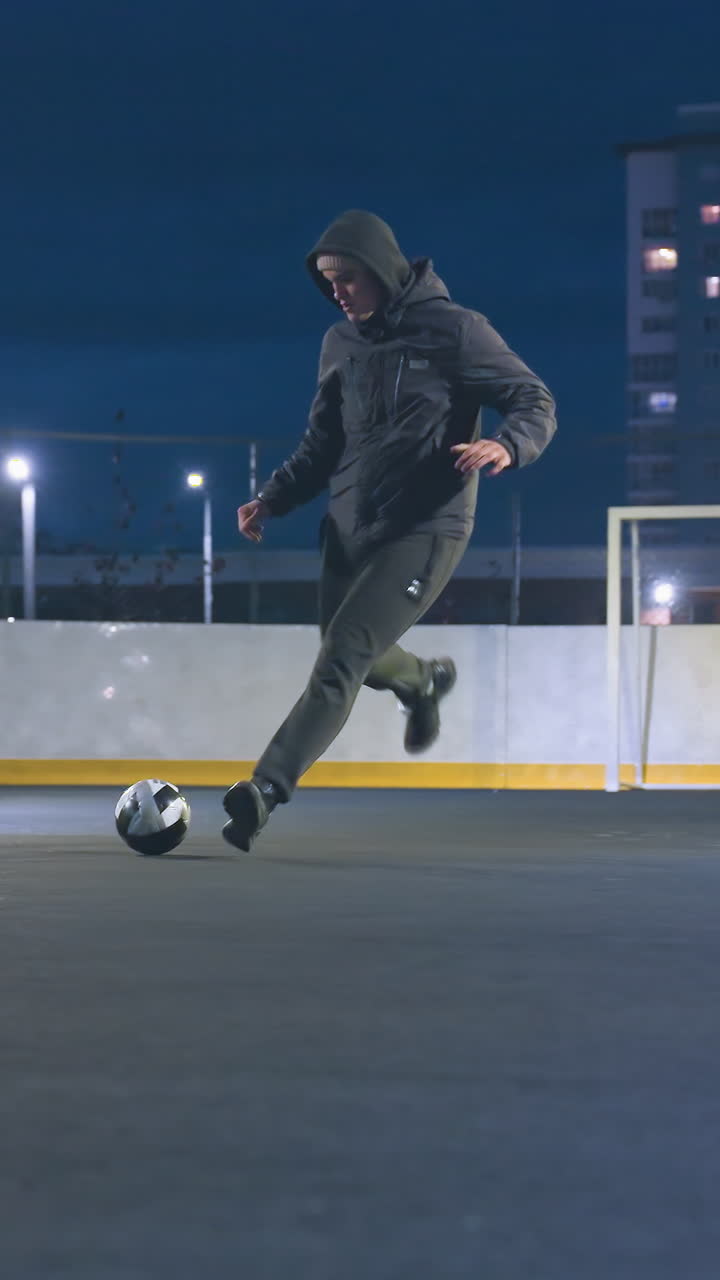 Player kicks soccer ball toward goal post during intense urban night practice, ball rolls out after hitting goal, surrounded by illuminated city buildings