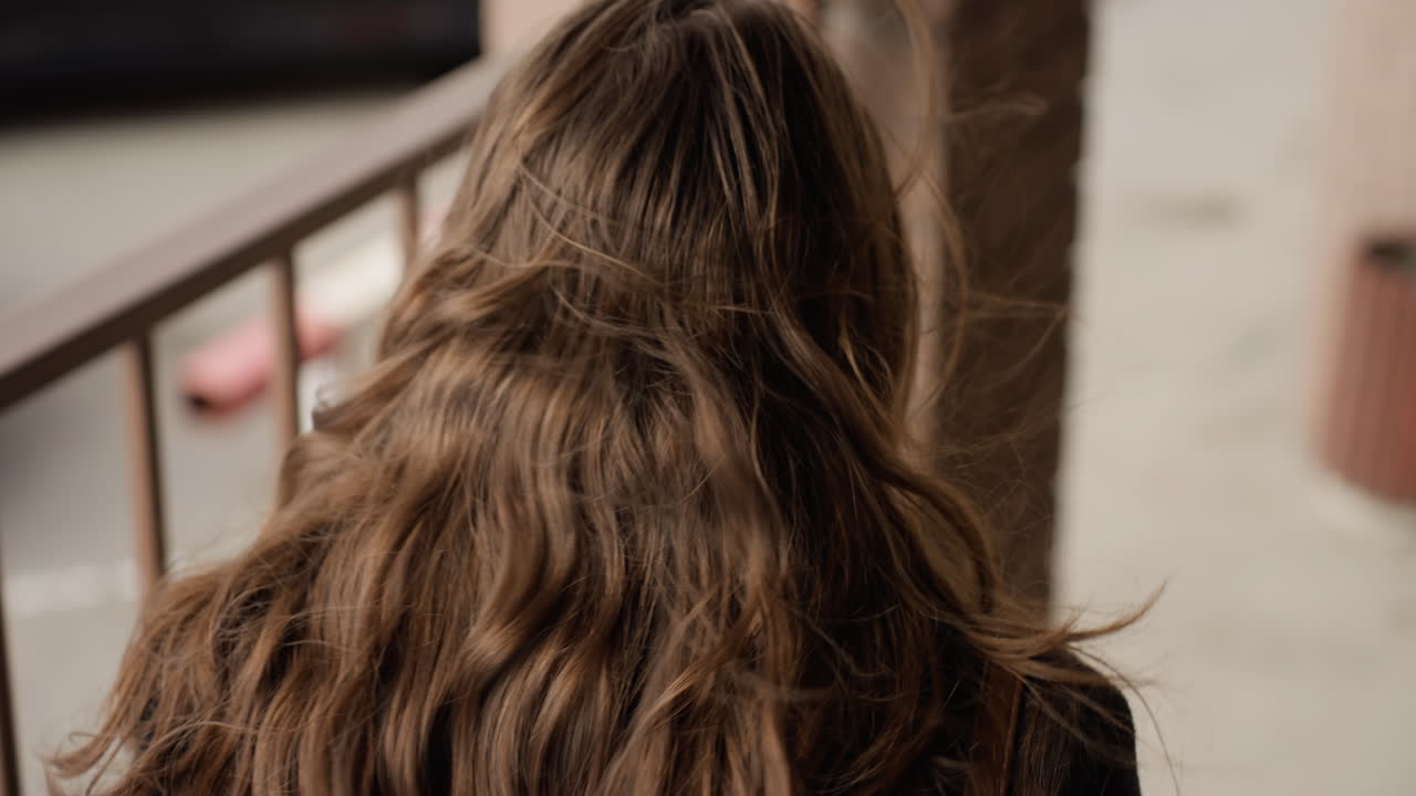 Vista aérea de una chica rubia que pasa junto a una pared de ladrillo, con una columna de hierro roja y blanca visible cerca del camino; la brisa levanta algunos mechones y la entrada urbana se percibe tranquila e iluminada por el sol durante un desplazamiento informal.