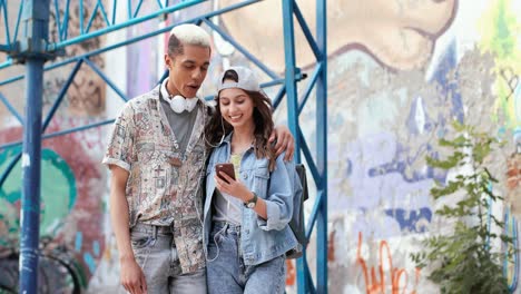 smiling man and his caucasian girlfriend wearing headphones and watching something on the smartphone while walking down the street