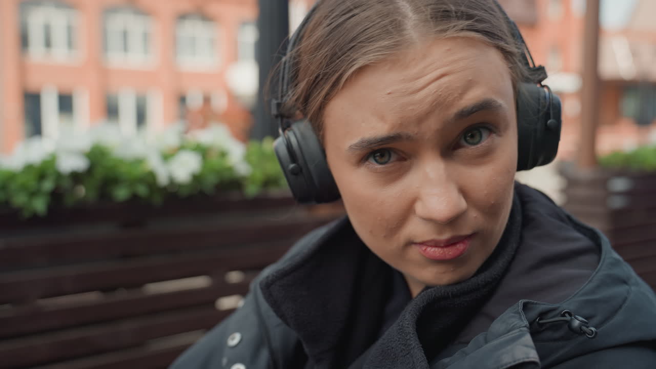 Giovane donna con cuffie che reagisce all'aperto su una panchina di una terrazza in un caffè urbano con edifici in mattoni e fioriere, indossa una giacca scura, sguardo scettico ed espressivo verso la fotocamera, leggera pioggerellina sui capelli