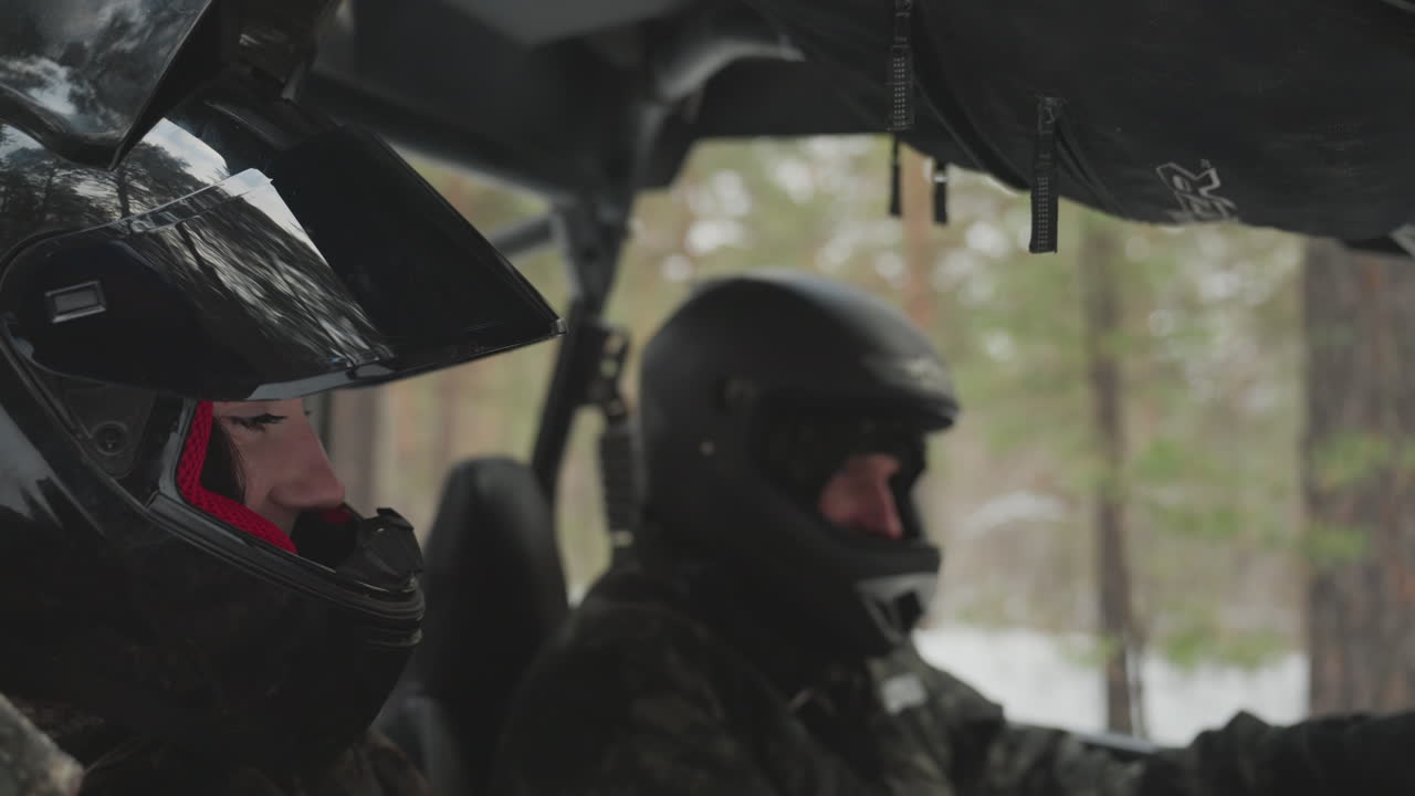 Two riders inside vehicle in snow forest, driver concentrates on steering while passenger checks route and instruments, helmets on, interior frosted, teamwork and safety emphasized