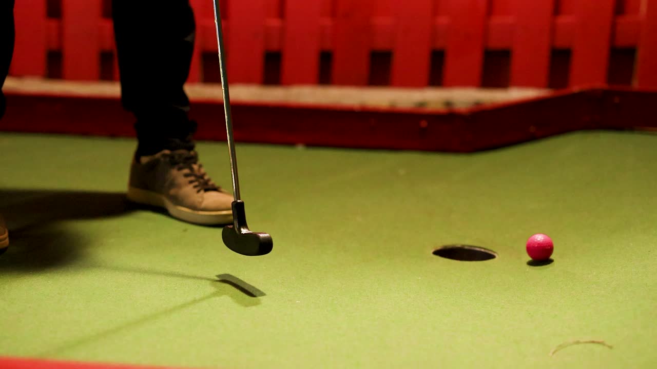 A player putts a pink ball towards a hole on a mini-golf course with colorful lighting