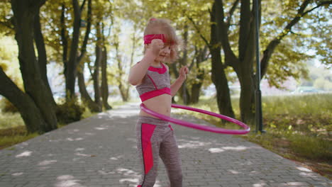 Athletic-fitness-children-girl-training-playing-twisting-Hula-hoop-circle-ring-around-waist-in-park