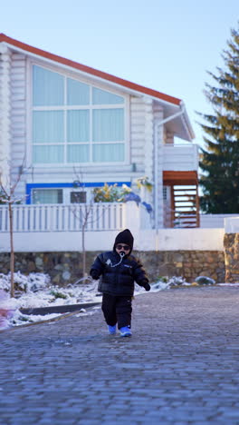 Lovely baby boy wearing warm black clothes and sunglasses approaches camera. Cute toddler kid with pacifier runs outdoors in winter. Vertical video.