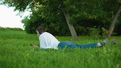 man lying on grassy field wearing headphones and white top with jeans, focused on phone, surrounded by vibrant greenery and trees, with people walking in blurred distant background