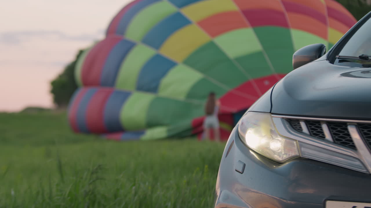 Close up front car bumper headlight framing blurred woman handling hot air balloon envelope on grassy field at dusk, colorful deflated balloon panels draped on ground under soft pastel sky