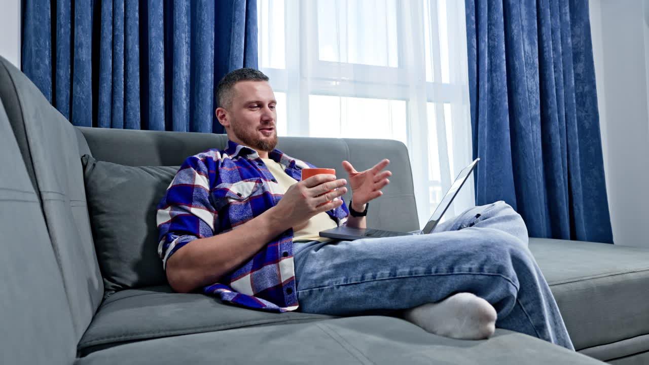 Mand sidder på sofaen med en drink i hånden, mens han bruger en bærbar computer i en moderne stue om dagen.