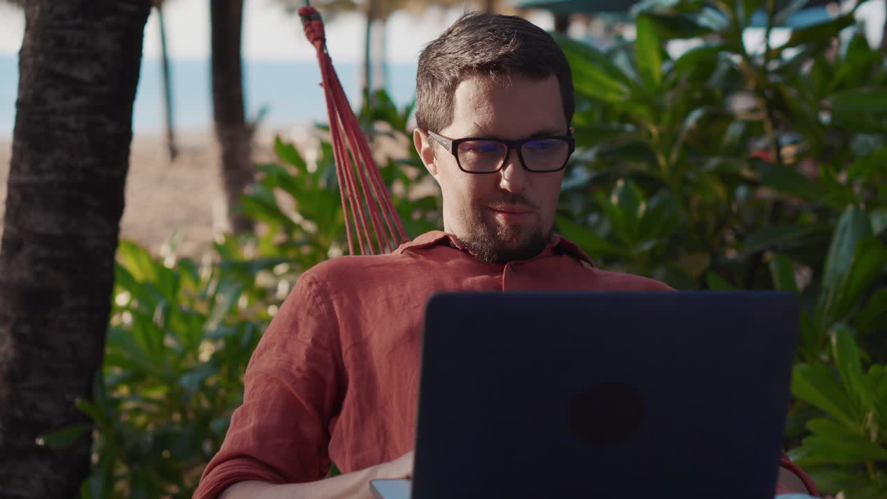 hombre trabajando en una computadora portátil en una hamaca en una playa tropical