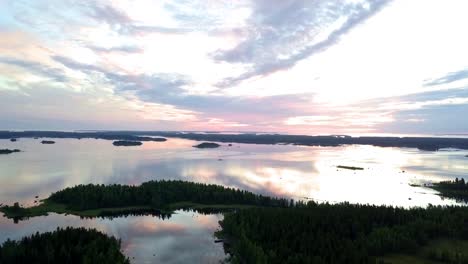 Colorful-Sunset-lighting-up-the-archipelago-with-vibrant-sky-reflected-like-a-mirror-in-the-calm-sea,-traditional-red-summer-cottage,-DRONE-SHOT