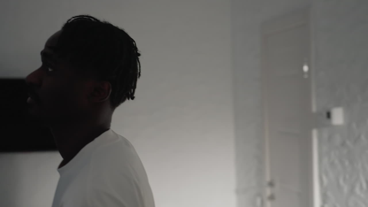 Man with dreadlocks wearing a white t-shirt bends over in a well-lit modern kitchen, focusing on something on the counter, with shadows cast on the walls and minimalist background design