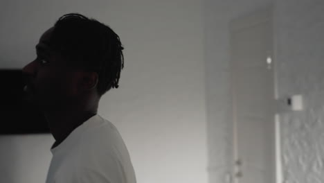 man with dreadlocks wearing a white t-shirt bends over in a well-lit modern kitchen, focusing on something on the counter, with shadows cast on the walls and minimalist background design