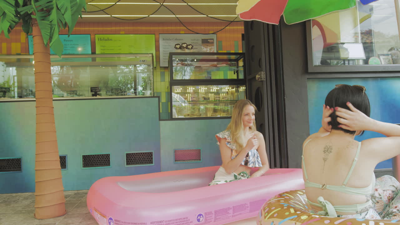 Two young women enjoy ice cream in a relaxed atmosphere next to an inflatable pool in front of a contemporary ice cream parlor.
