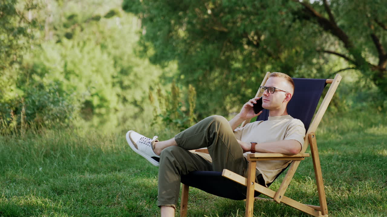 Serious Caucasian male sits in a folding chair having phone conversation. Man combining work and rest in nature.