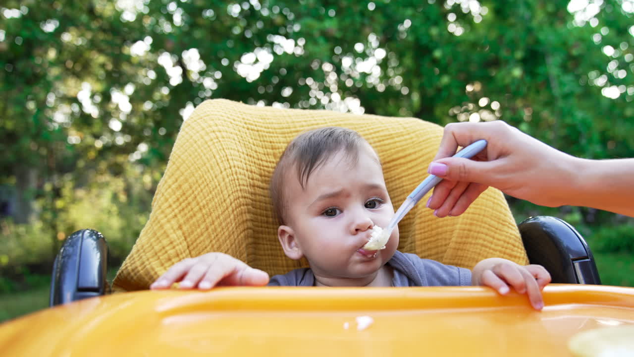 Adorable baby boy sitting in yellow chair outdoors. Mother gives full spoon to her child and he eats. Blurred nature backdrop.