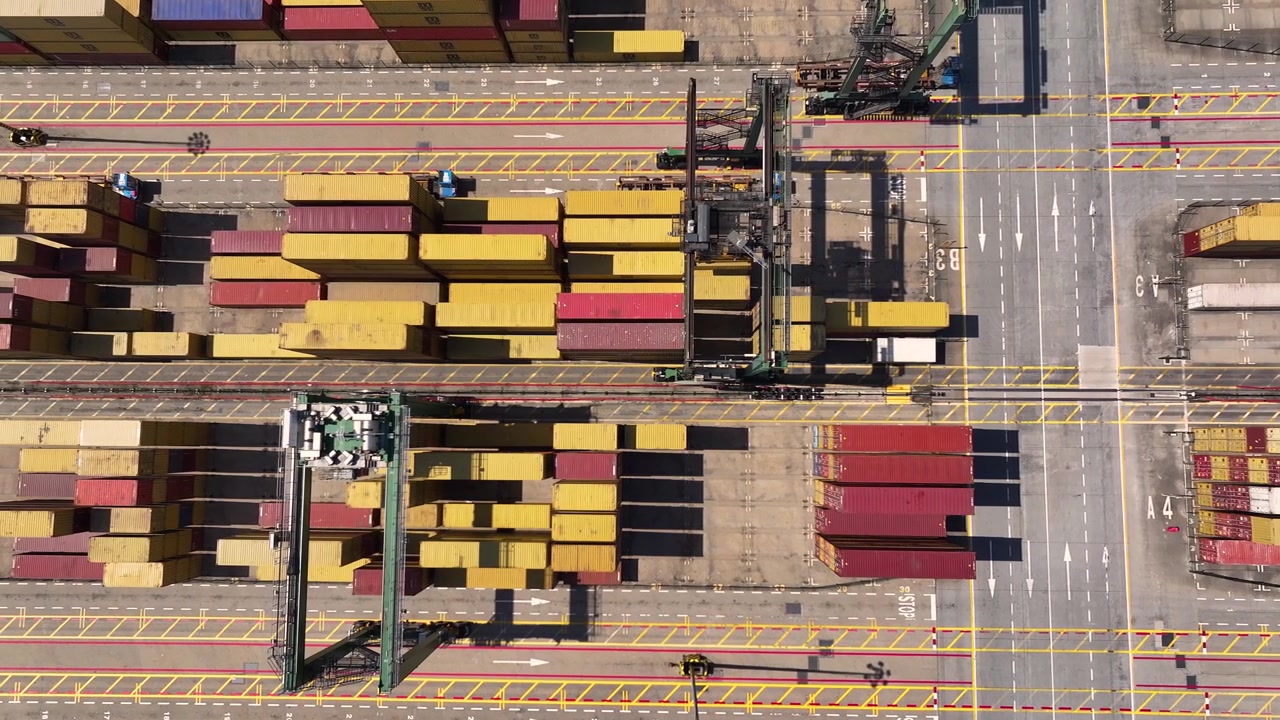 Container yard shows numerous stacked cargo containers. Cranes operate to load and unload freight. Port area is busy with shipping activities. Delays in delivery are visible.