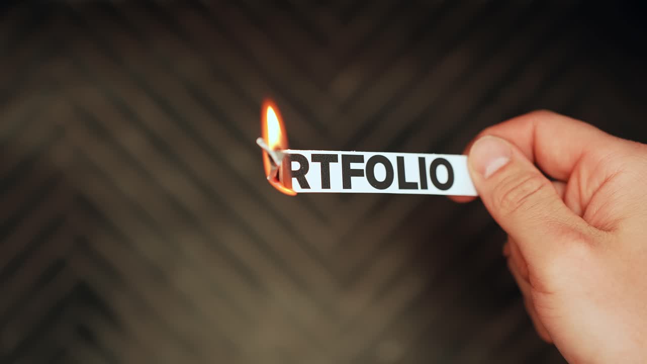 Hand burns a paper label reading PORTFOLIO, symbolizing investment risk, market losses, and financial uncertainty