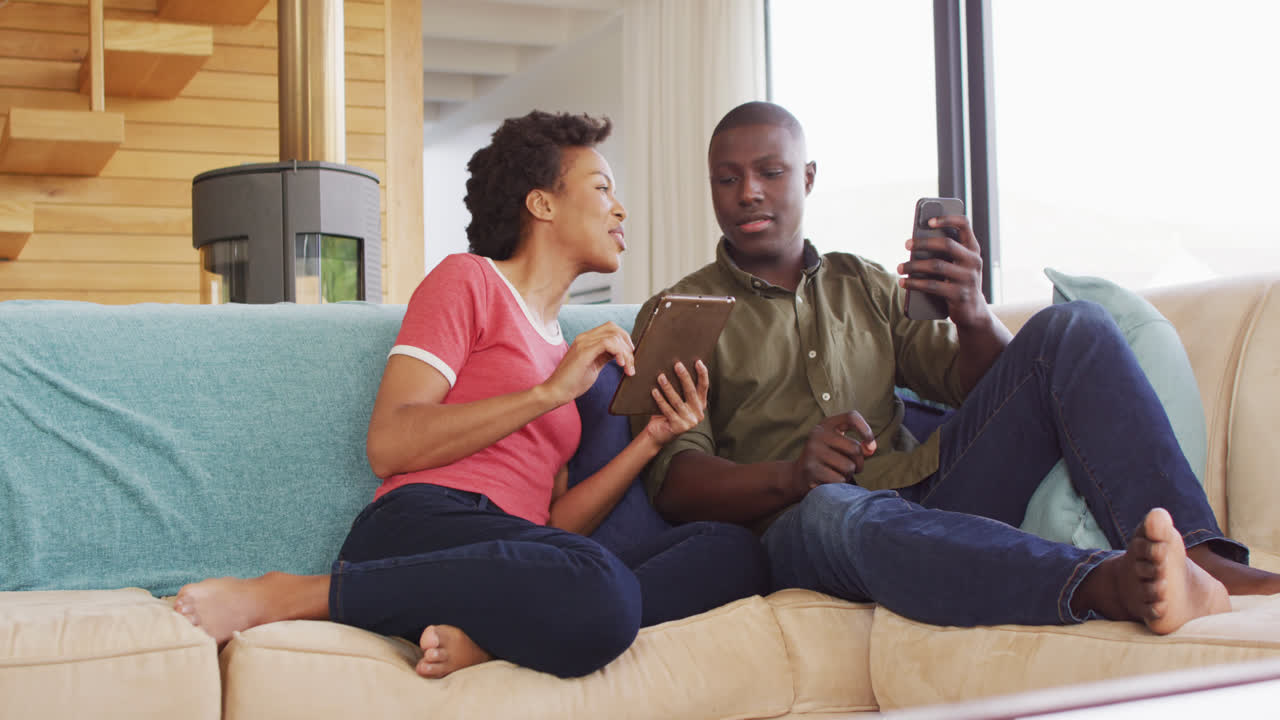 Video of happy african american couple sitting on sofa and using tablet