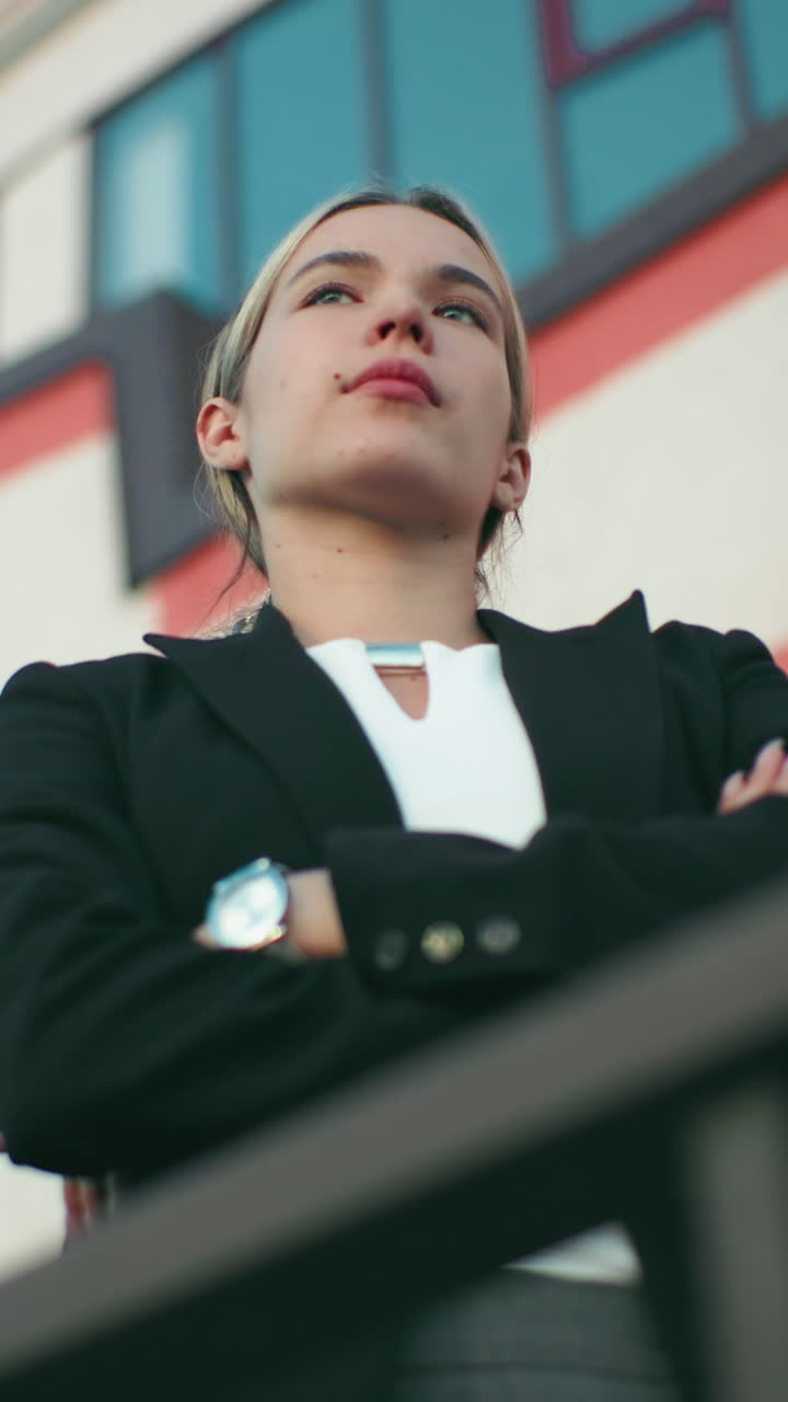 Woman in black blazer and white top stands confidently with arms folded, gazing into distant sky in front of modern building, exuding ambition