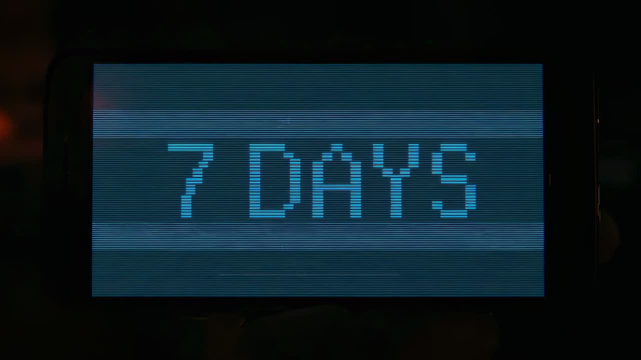 Displaying handheld display showing pixelated cyan 7 DAYS on power-up in dim room, bezel banding