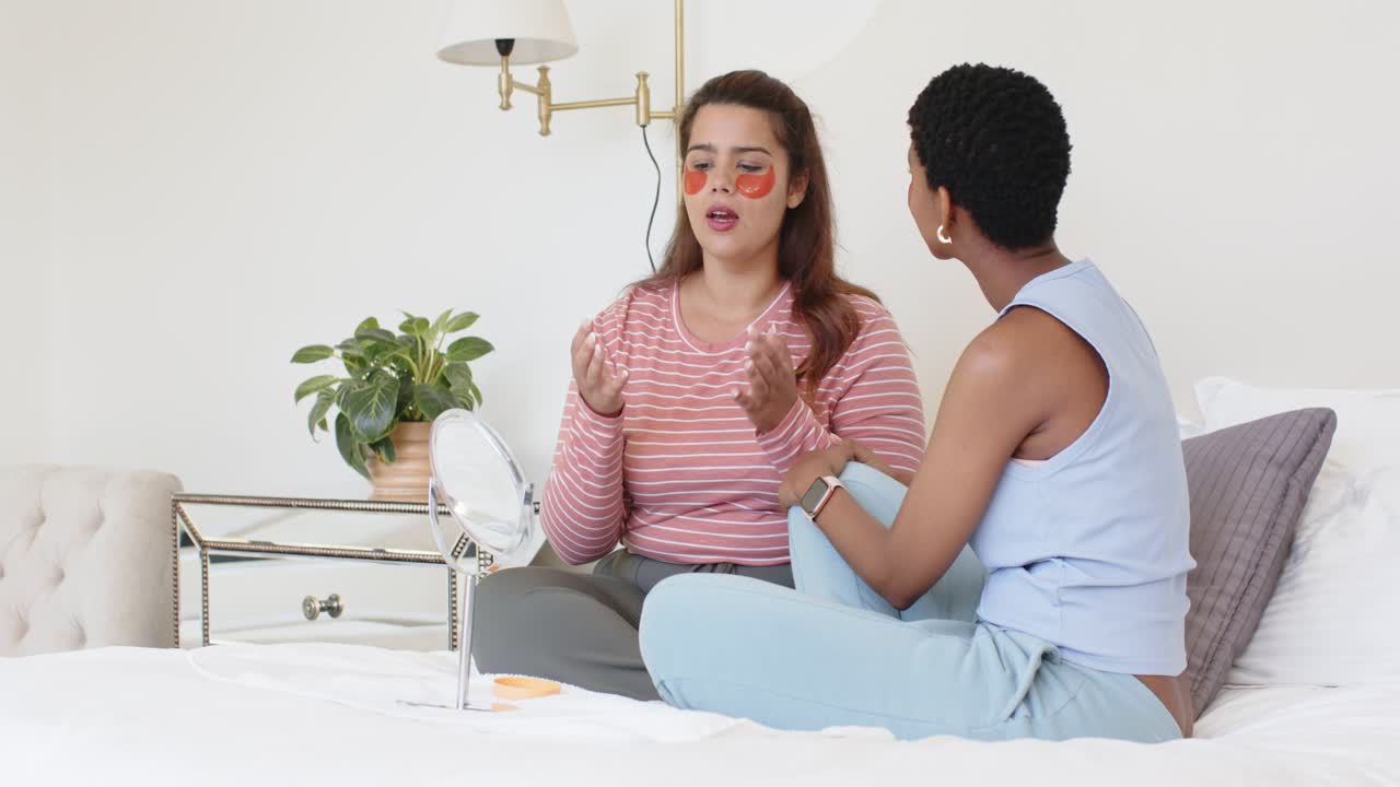Two women placing orange under-eye patches, massaging face on bed using mirror and jar, teaching