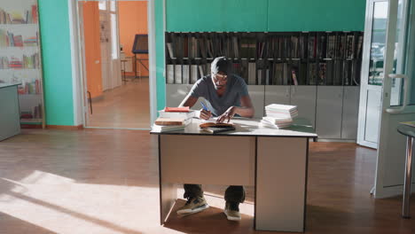 high school student sits at desk in bright library, writing in notebook with pen while moving legs, surrounded by books, deeply focused on studies as sunlight streams across floor