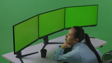 vista lateral de una mujer asiática aburrida trabajando con múltiples monitores de computadora en un estudio de pantalla verde