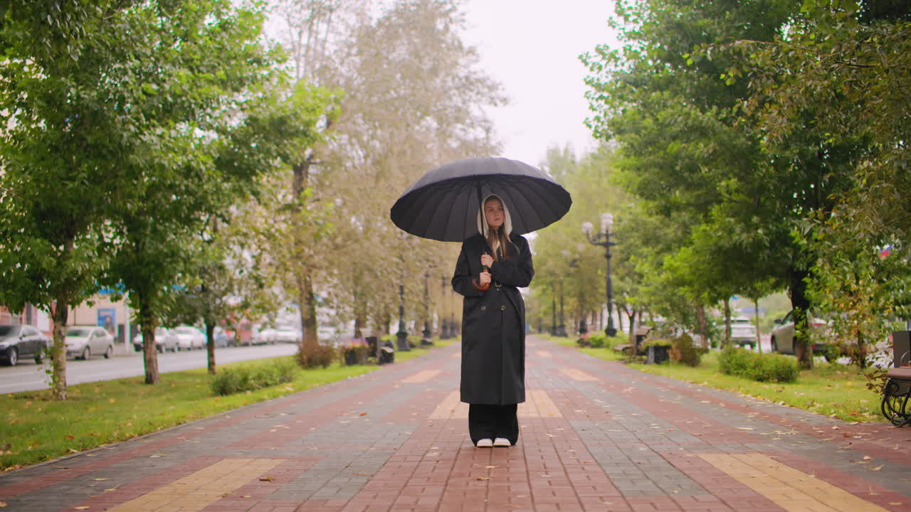 Woman in long black trench coat holding umbrella standing on wet park walkway during rainy autumn day, surrounded by trees, cars, benches, cloudy sky, urban outdoor scene with reflective calm mood