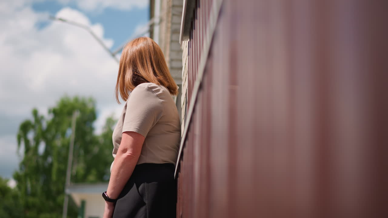 Tired lady walking toward brick building under warm summer sunlight, holding bag and stopping to rest against wall, expressing exhaustion, quiet thought, and emotional pause