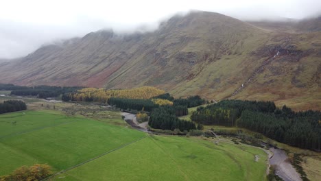 Aerial-view-of-green-hills-and-river-in-Scottish-highlands-with-trees-and-yellow-leaves,-misty-and-cloudy