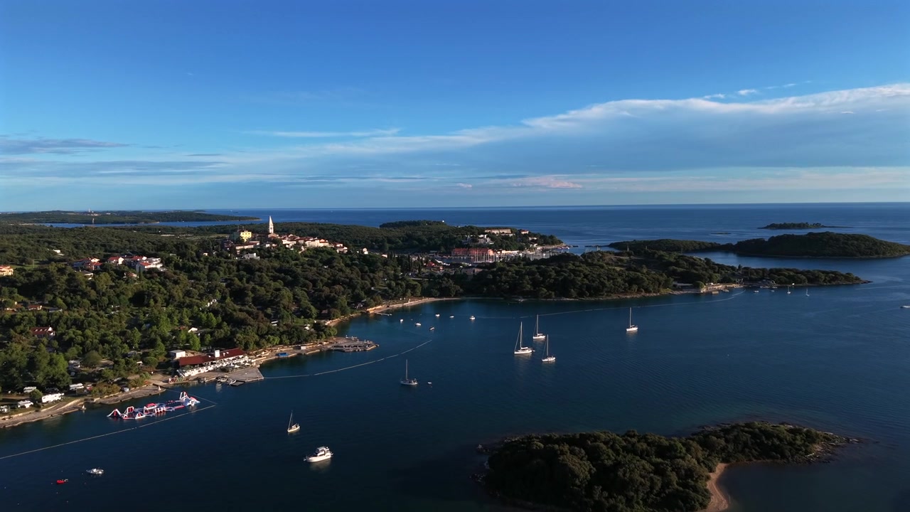 Isole boscose punteggiano la scintillante baia adriatica di un blu profondo lungo la costa istriana in Croazia, con un colorato parco acquatico gonfiabile che galleggia vicino a una spiaggia sabbiosa in un resort di campeggio estivo. Ripresa aerea con drone in avvicinamento.