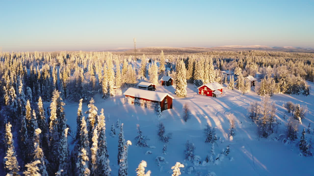 Luftfoto over soloppgang som skinner på snødekte røde vinterhytter, omgitt av rimfrostkledde skogtrær