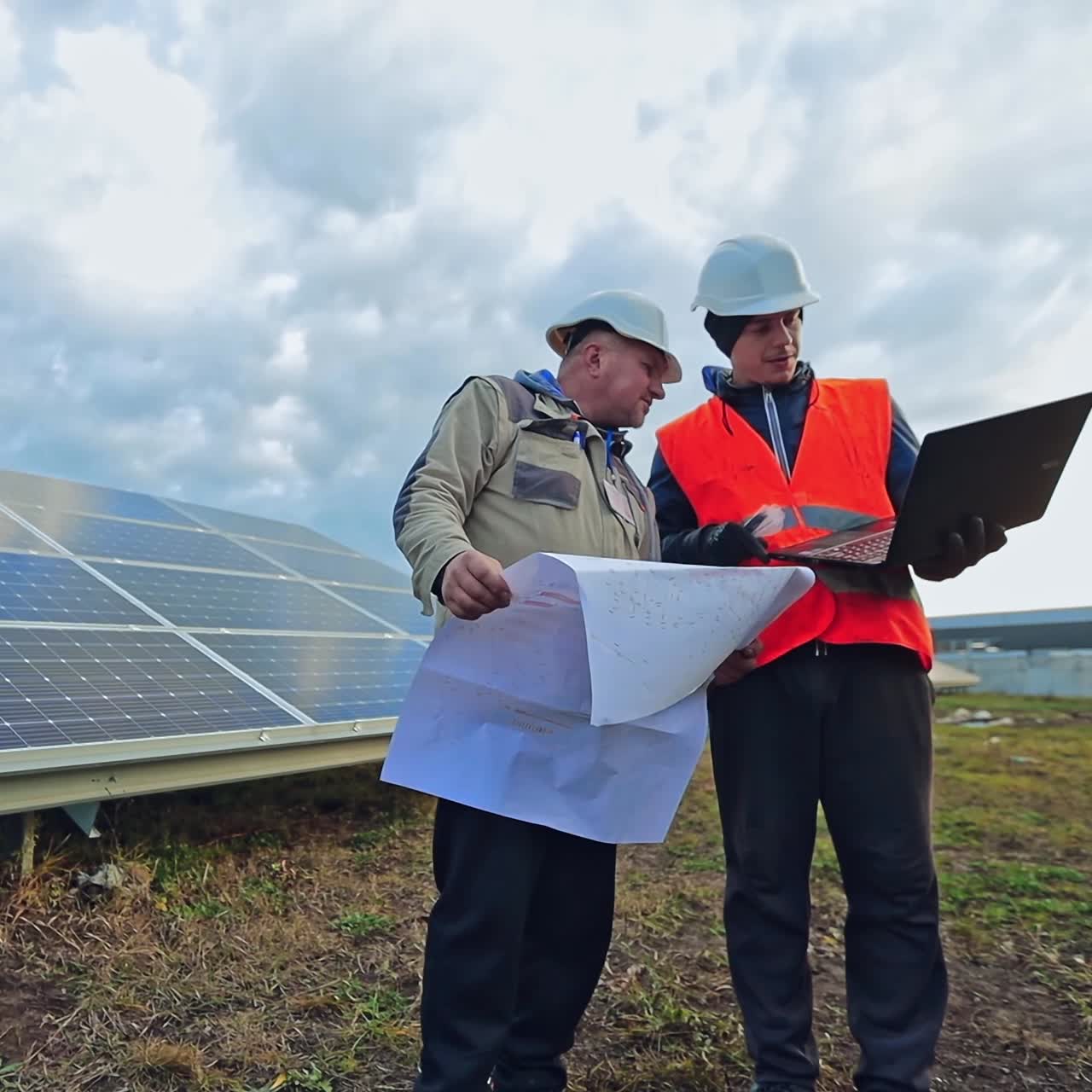 Men in helmets talking about solar panels. Managers discussing the plan on the field with solar panels using a computer laptop.