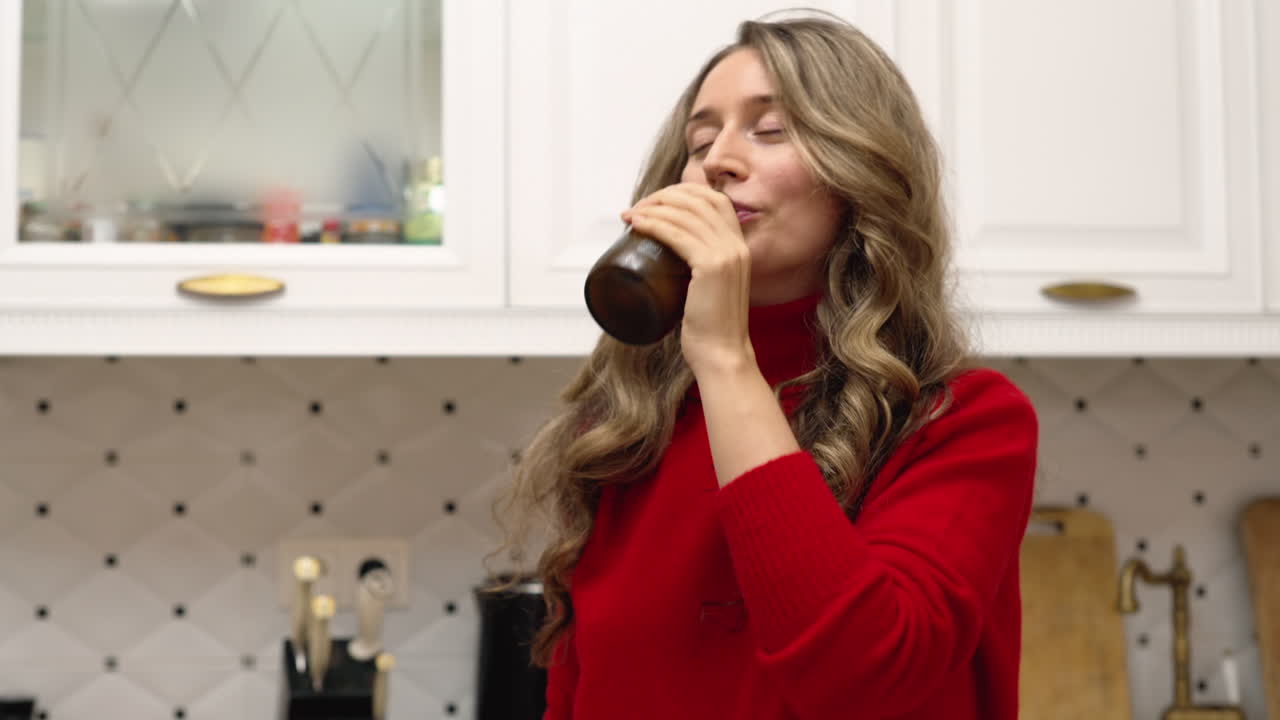 Woman drinking kombucha from a brown glass bottle in the kitchen
