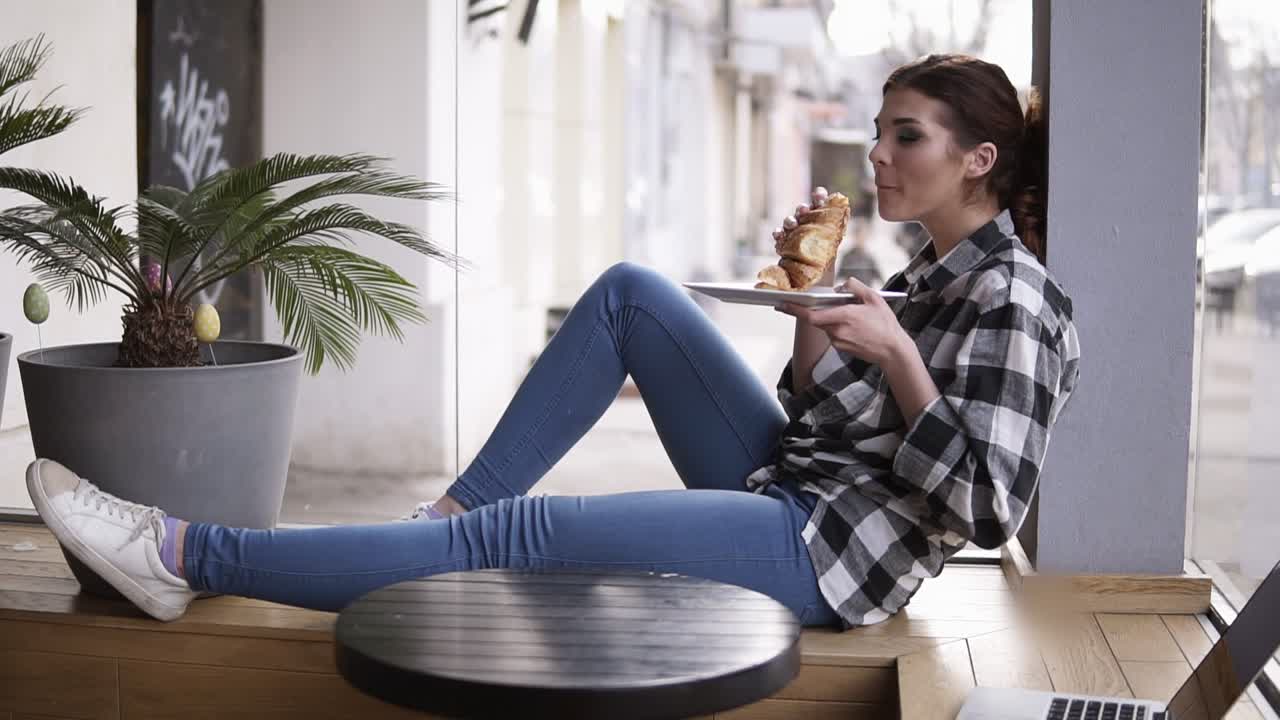 vista lateral de una chica joven y bonita sentada en una cafetería en un asiento de madera junto a las ventanas brillantes. la chica está comiendo un croissant. mastica y cierra los ojos. disfruta del tiempo