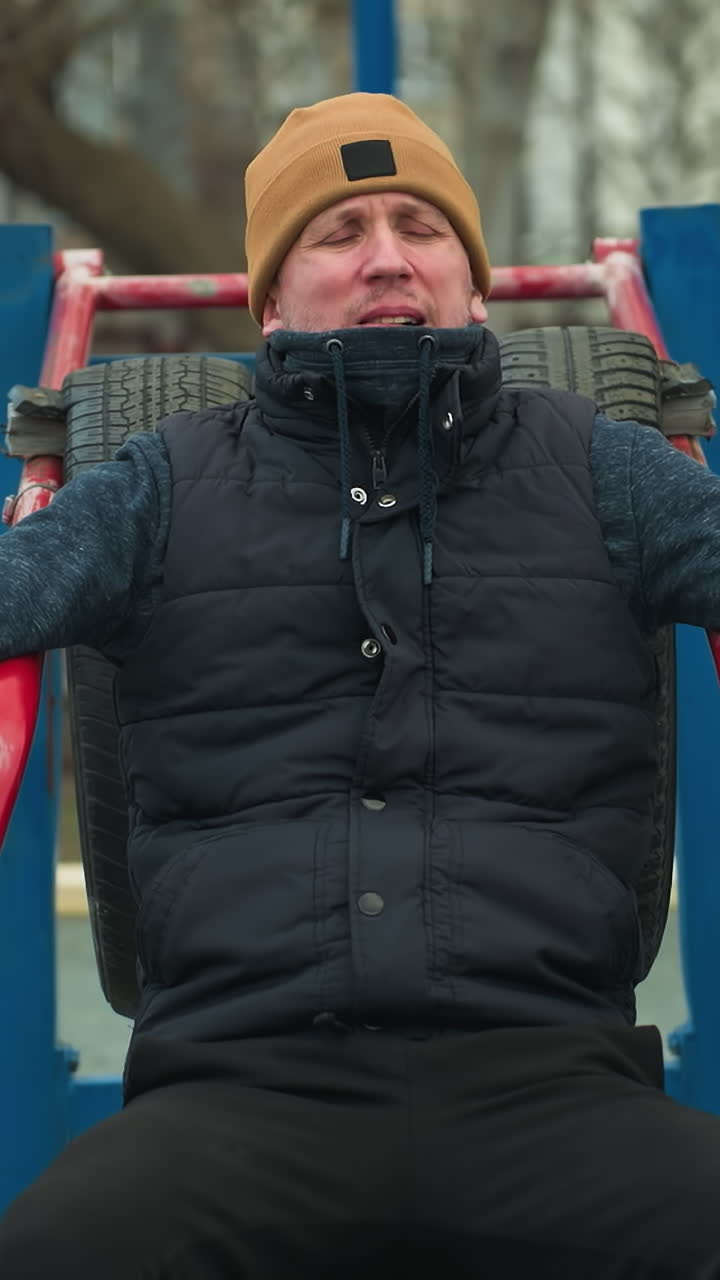 A coach seated on workout equipment, adjusting his beanie, looks tired, in the background, a boy dressed in black plays football