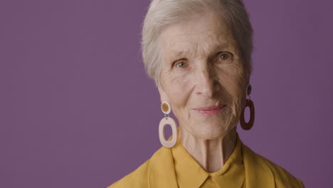 senior woman with short hair wearing mustard colored shirt and jacket and earrings posing and smiling at camera on purple background