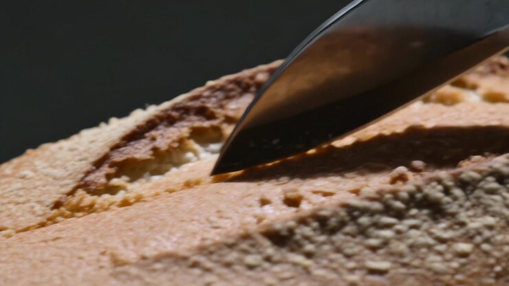 Slicing golden-brown crust loaf with metal blade on studio tabletop, revealing pale crumb fragments