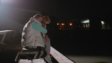 dog owner rests her head affectionately on back of seated dog during peaceful night scene on wooden bench under bright outdoor lights, showing tender bond of love, and deep emotional connection
