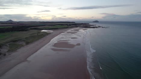 imágenes rápidas de drones aéreos que vuelan alto sobre una larga playa de arena al atardecer mirando a través del océano ondulante mientras la marea golpea suavemente la orilla