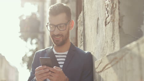 el primer plano de un hombre sonriente vestido con estilo apoyado contra la pared en la calle y enviando mensajes de texto al teléfono inteligente