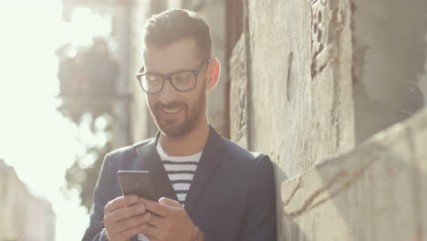 close up of a smiling man in stylish outfit leaning against the wall in the street and texting on the smartphone