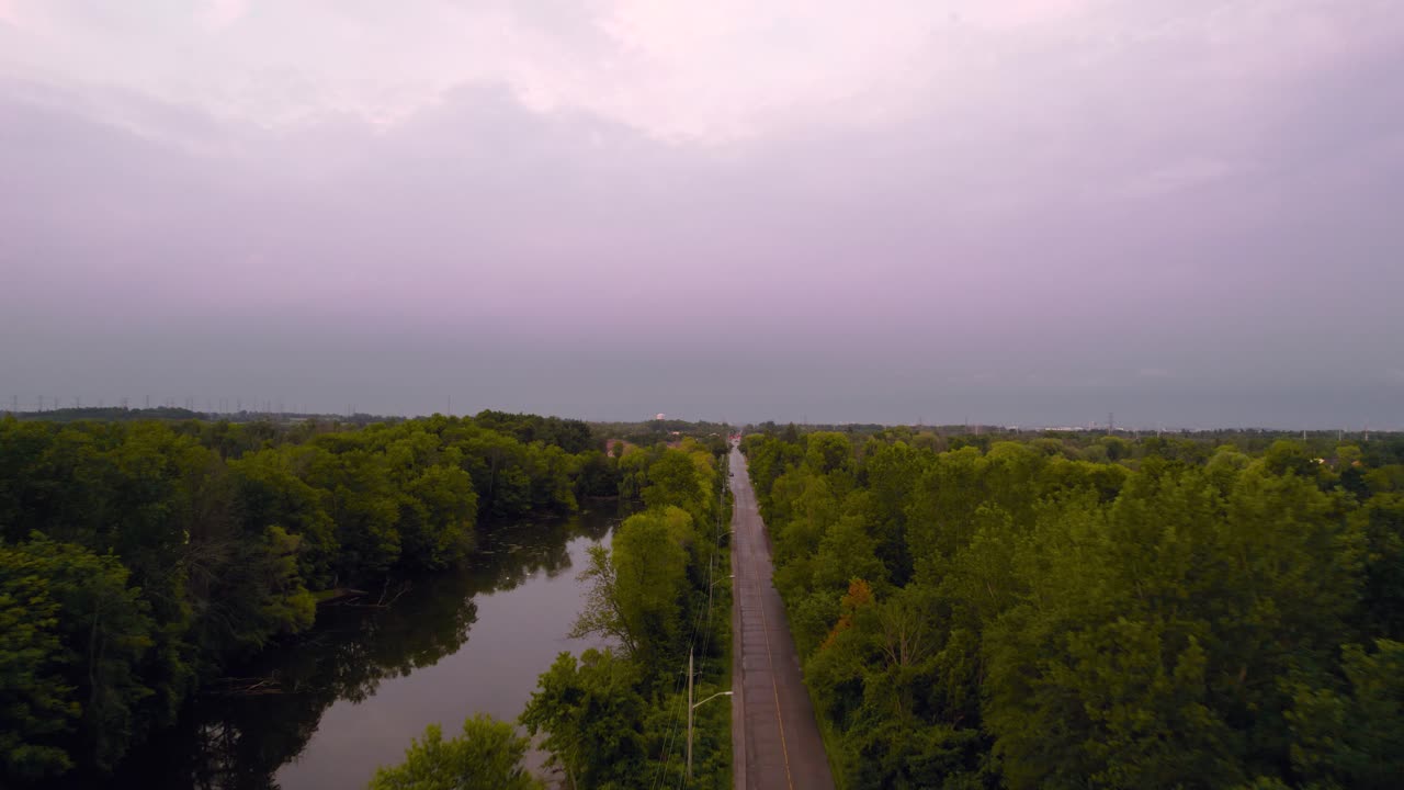 toma aérea de seguimiento de drones de la carretera rural al atardecer junto al estanque y la zona boscosa con un paisaje escénico de cielos nublados